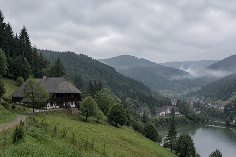 Dark fir-covered hills above a Black Forest lake, a small village below and mist in the valley.