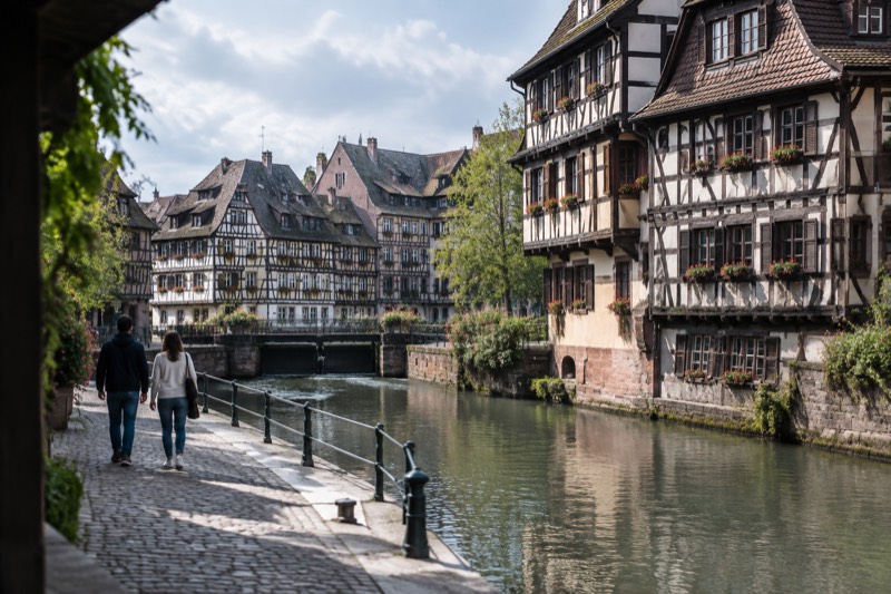 Two walkers on a stone quay beside the river, half-timbered houses across the water, seen from behind.