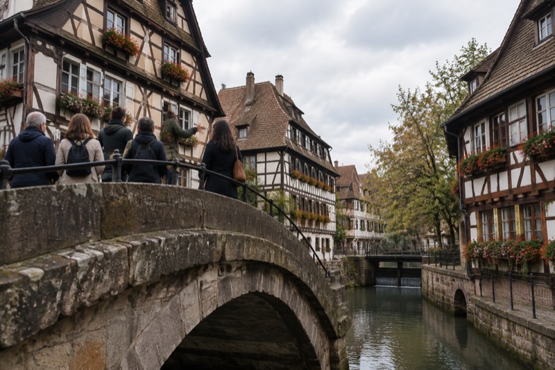 A small group on a stone bridge listening to a guide, seen from behind, canal houses in the background.