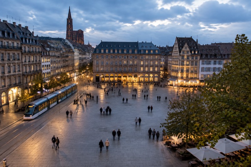 A lit evening square in central Strasbourg, warm shop windows and a tram gliding past, blue-hour sky