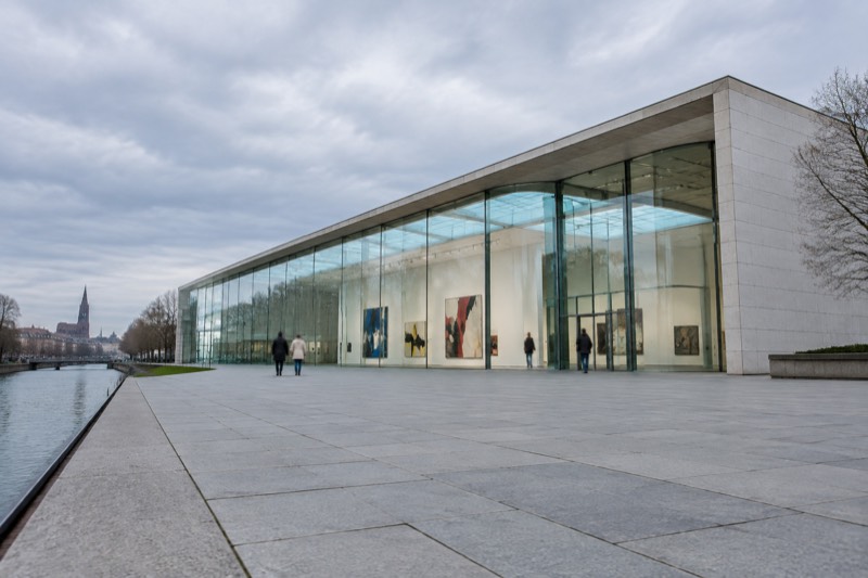A bright modern art museum interior with a tall white gallery and large abstract paintings, glass roof light