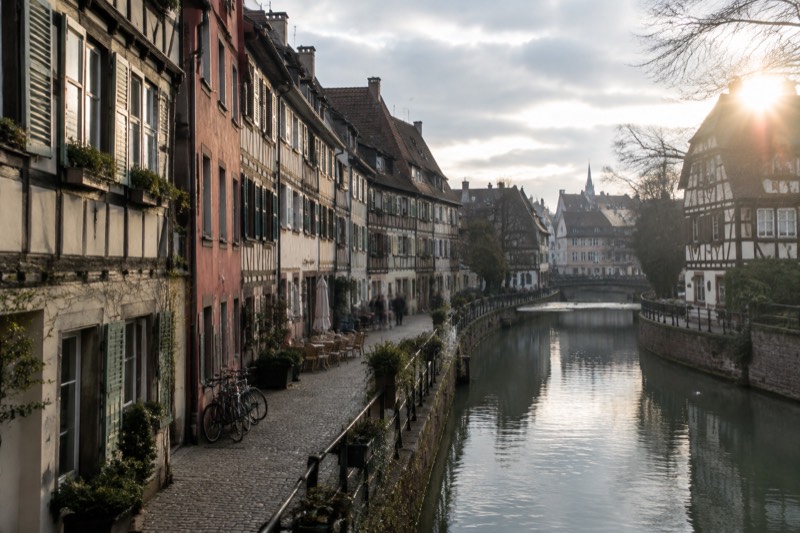 A calm canal-side street with narrow houses, bicycles against a wall and a few café chairs.