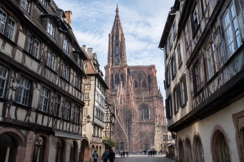 Pink sandstone cathedral facade with one tall spire seen along a narrow street in morning light