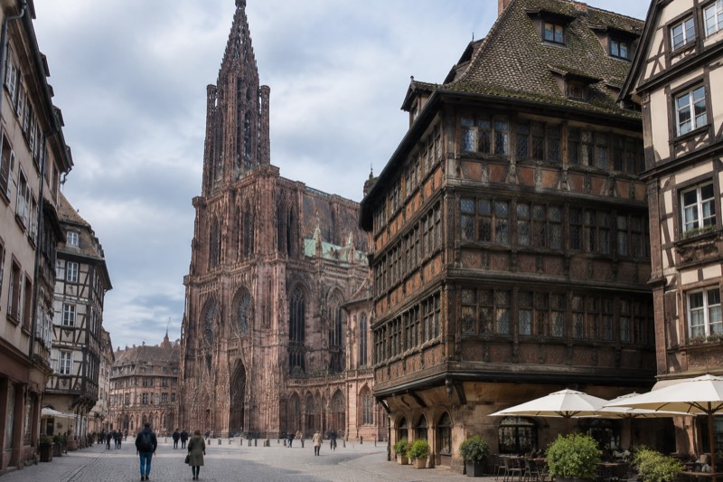 Richly carved half-timbered merchant house beside a cathedral square at lunchtime, distant figures crossing cobbles