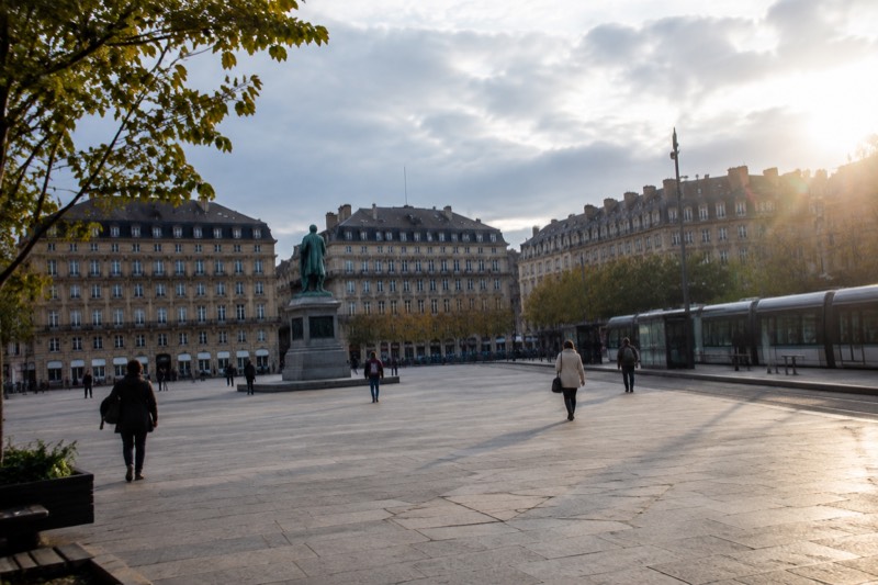 Wide open square with statue, tram stop and tall buildings on a bright day, people crossing.