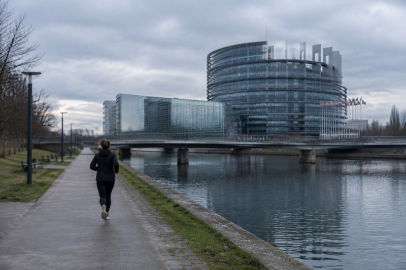 Curved glass and steel parliament building beside a canal with flags out of focus under blue sky.