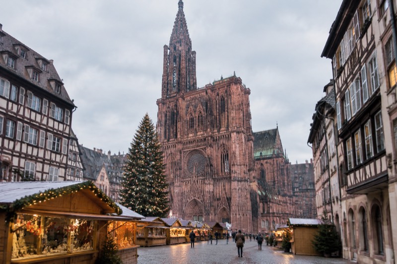 Wooden Christmas market chalets with warm lights in front of the cathedral at dusk, a large decorated fir tree