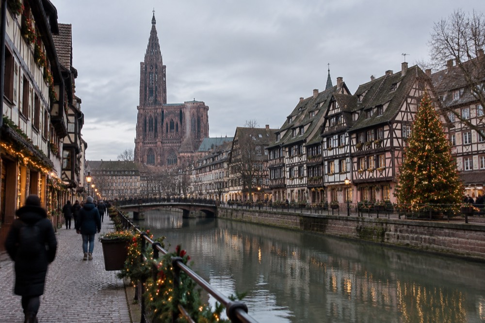 Festively decorated half-timbered buildings and a lit Christmas tree beside a canal in Strasbourg at dusk