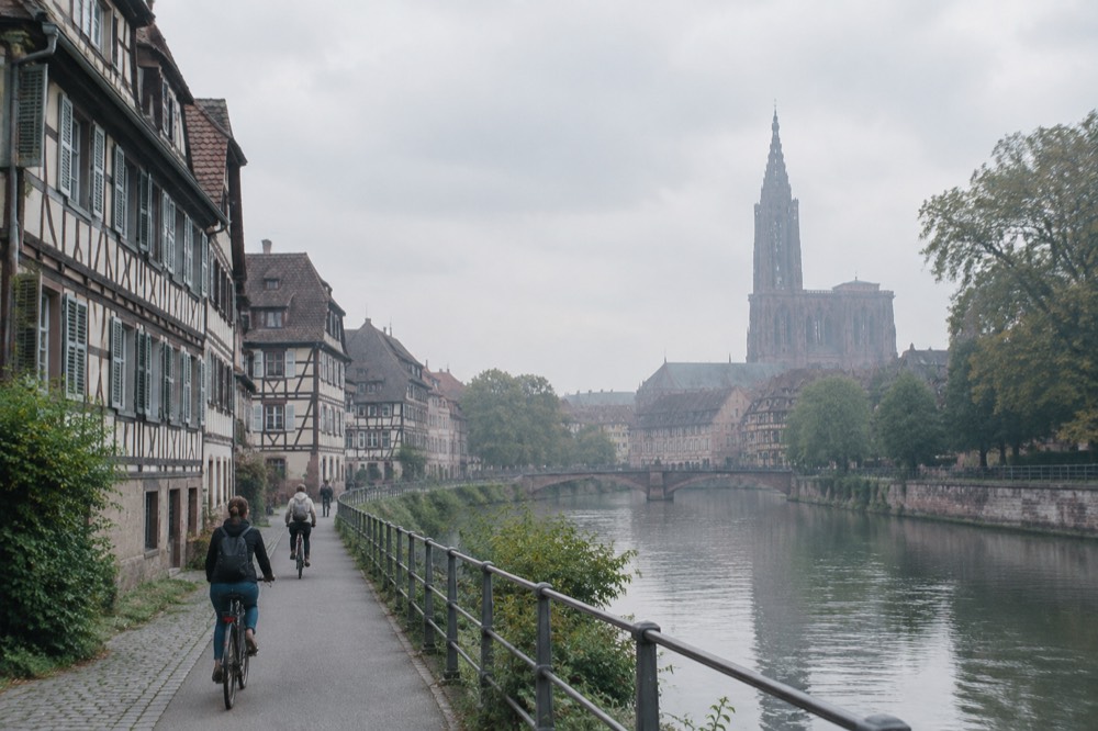 Cyclists on a riverside path beside the Ill with the spire of Strasbourg Cathedral visible in the distance