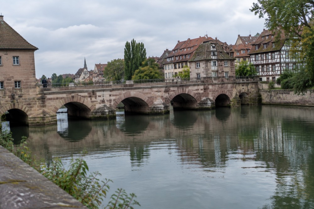 Stone arches of a historic barrage across a river with half-timbered houses reflected in the water