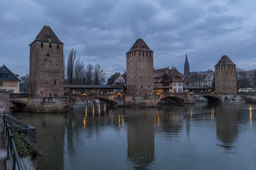 Covered bridges with square stone towers seen across a calm river at dusk