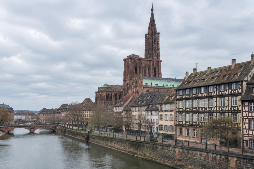 A Gothic cathedral spire rising above old rooftops seen from a riverside street