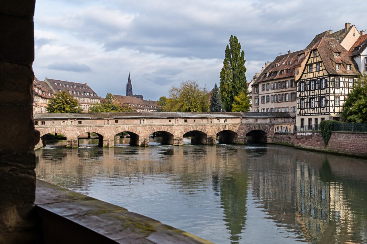 Stone arches of a historic barrage across a river with half-timbered houses reflected in the water