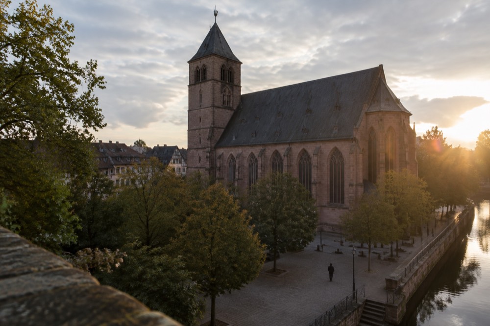 A stone square with trees and the side wall of an old church in evening light