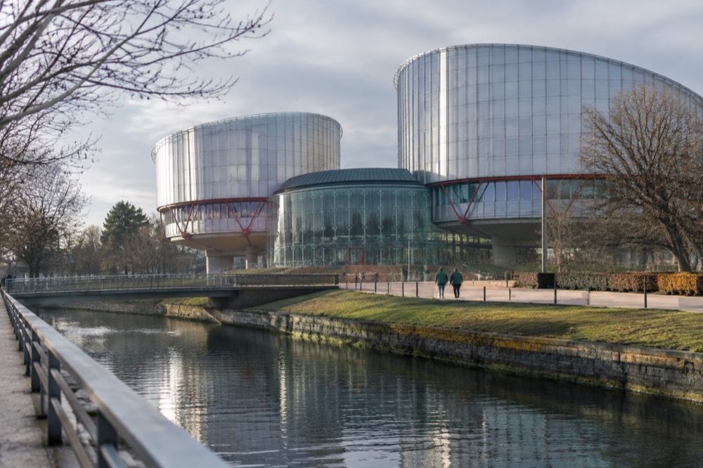 The glass and steel facade of a modern European court building beside trees and a wide plaza