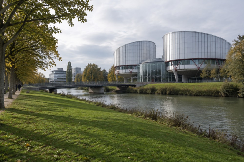 A wide lawn with modern institutional buildings and mature trees along a riverbank path
