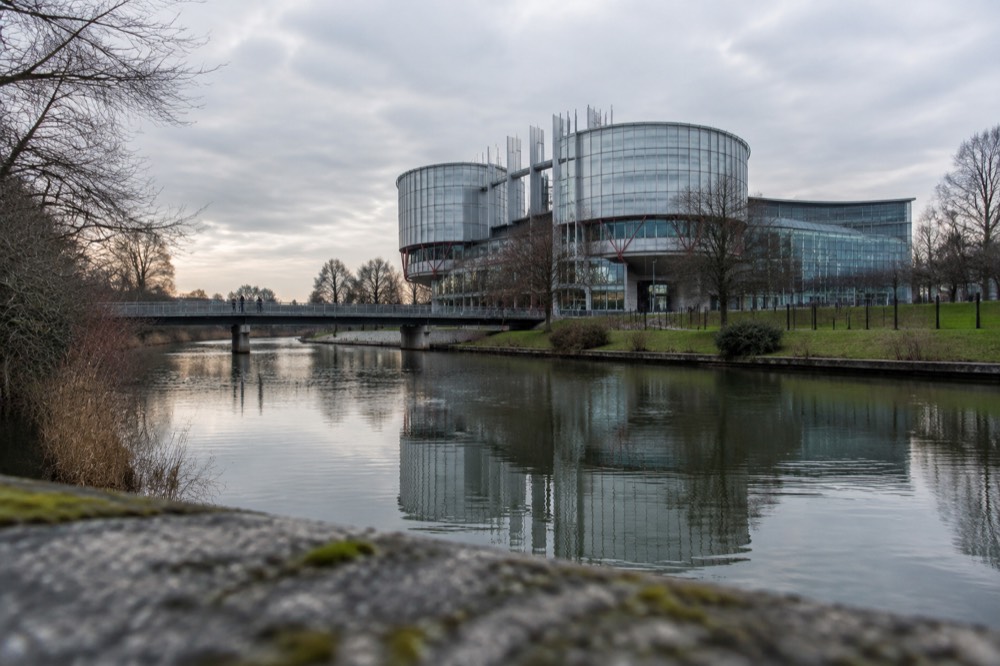 A quiet river with trees on both banks and the glass facade of an institutional building reflected in the water
