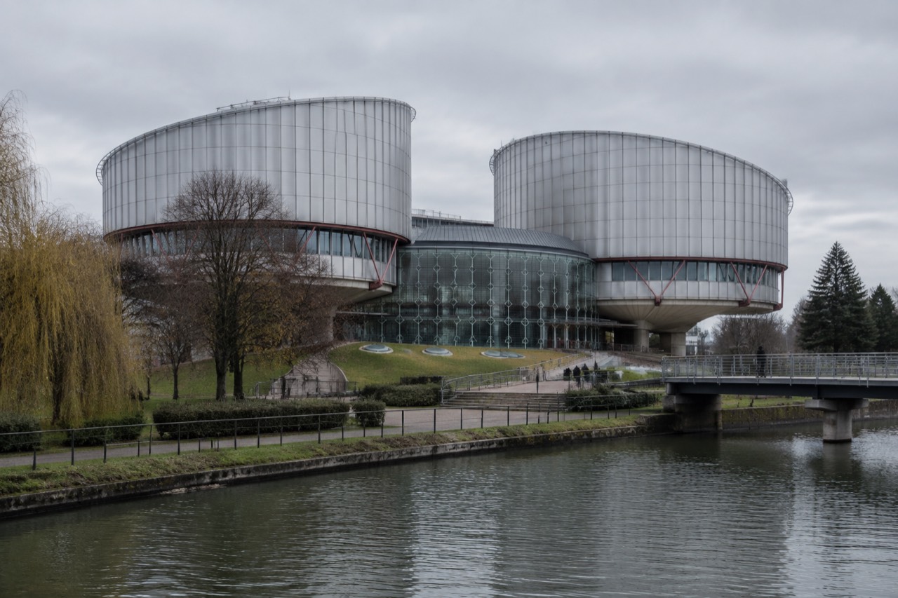 The glass and steel facade of a modern European court building beside trees and a wide plaza