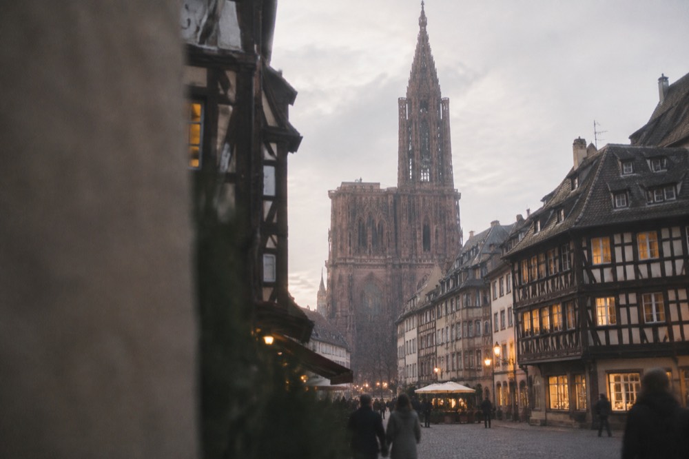 Old square at dusk with lit windows on timbered houses and a cathedral tower above