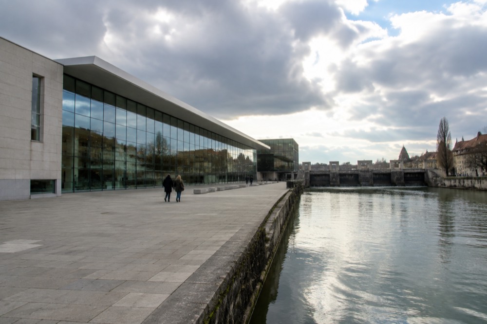 A modern glass-fronted museum building beside a calm river with a stone dam in the background