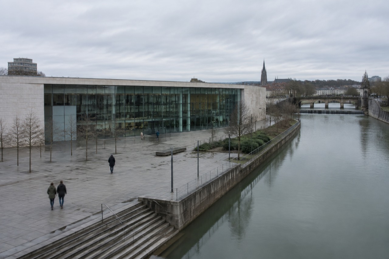 A modern glass-fronted museum building beside a calm river with a stone dam in the background