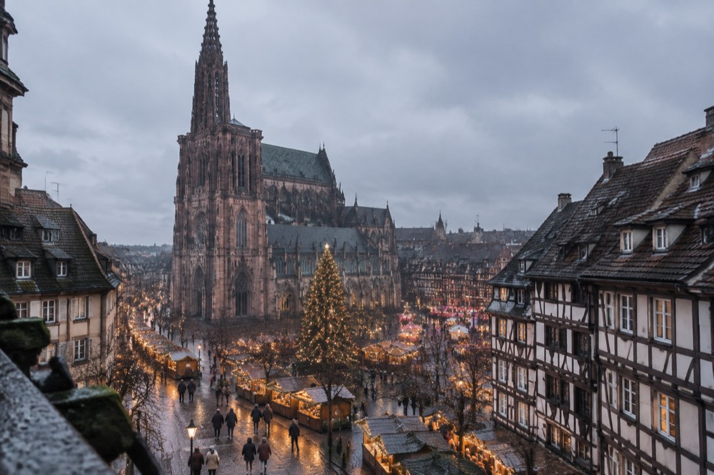 Winter street scene in Strasbourg with a decorated Christmas tree above the rooftops and market lights along a cold evening street.