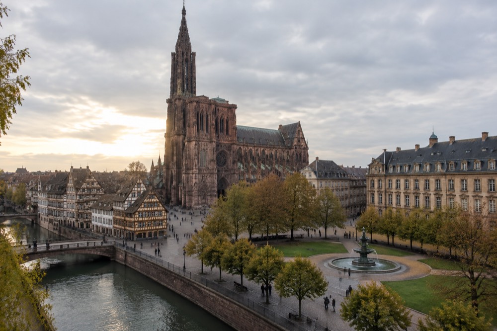 A broad tree-lined square in Strasbourg with a large fountain, lawns and stately stone buildings on three sides