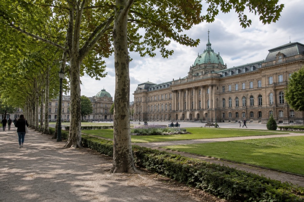A long classical building with columns beside a square, seen from a tree-lined pavement