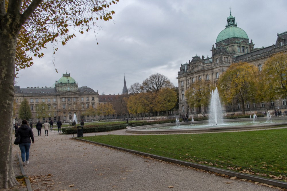 Fountains and lawns in a city square with autumn trees and grand institutional buildings behind