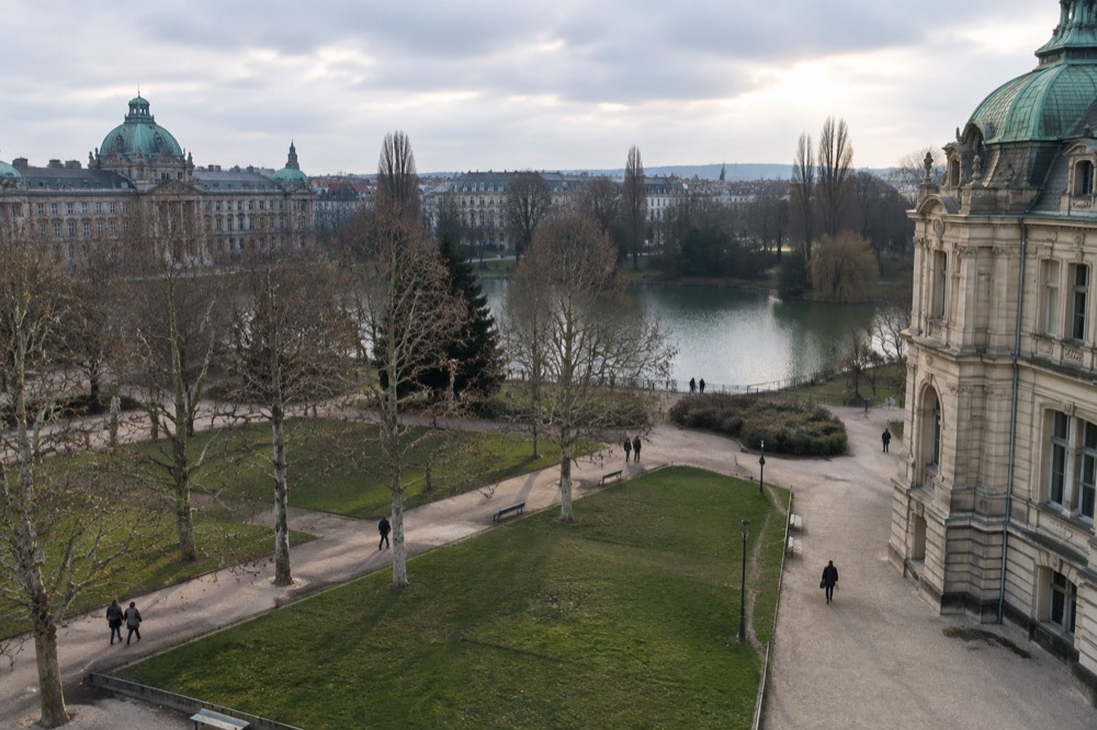 A city park with a lake, tall trees and stately buildings in the background