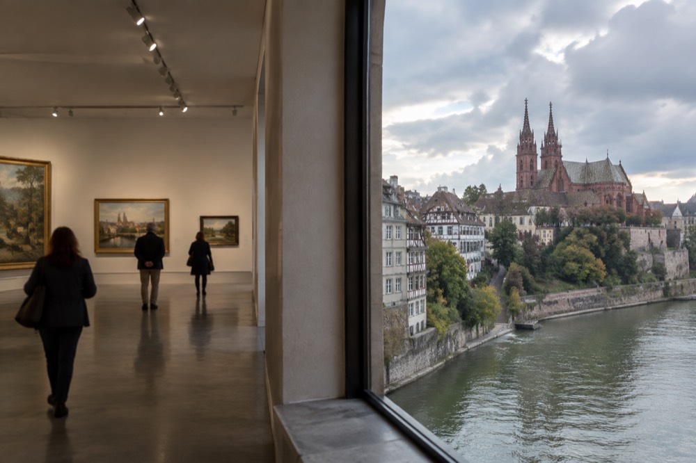 A gallery hall with paintings hung on pale walls and visitors walking between them