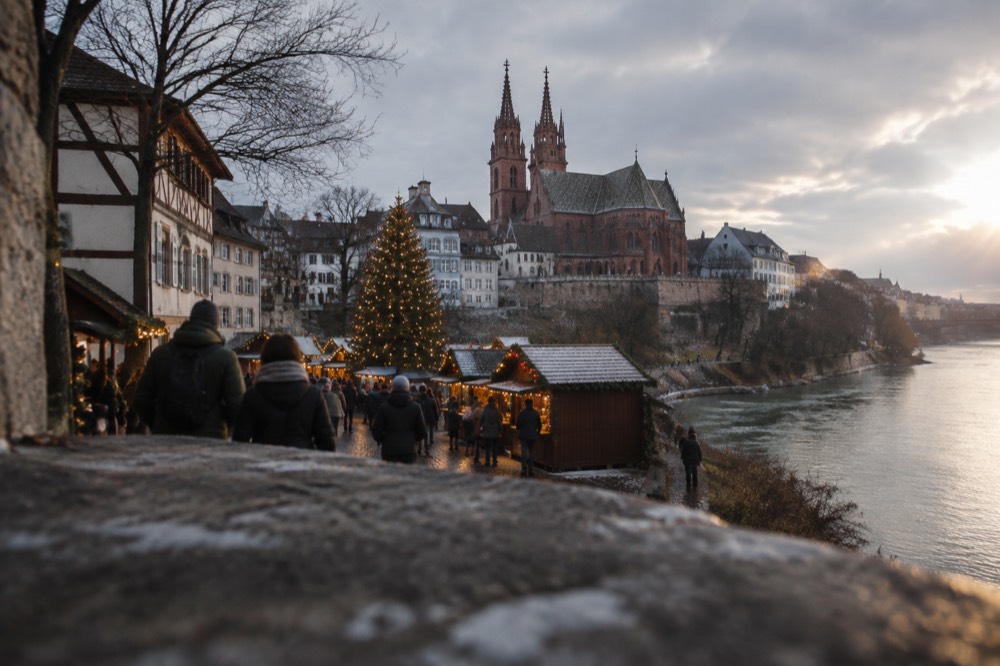 A Christmas market square with a tall decorated tree and wooden chalets glowing in early evening