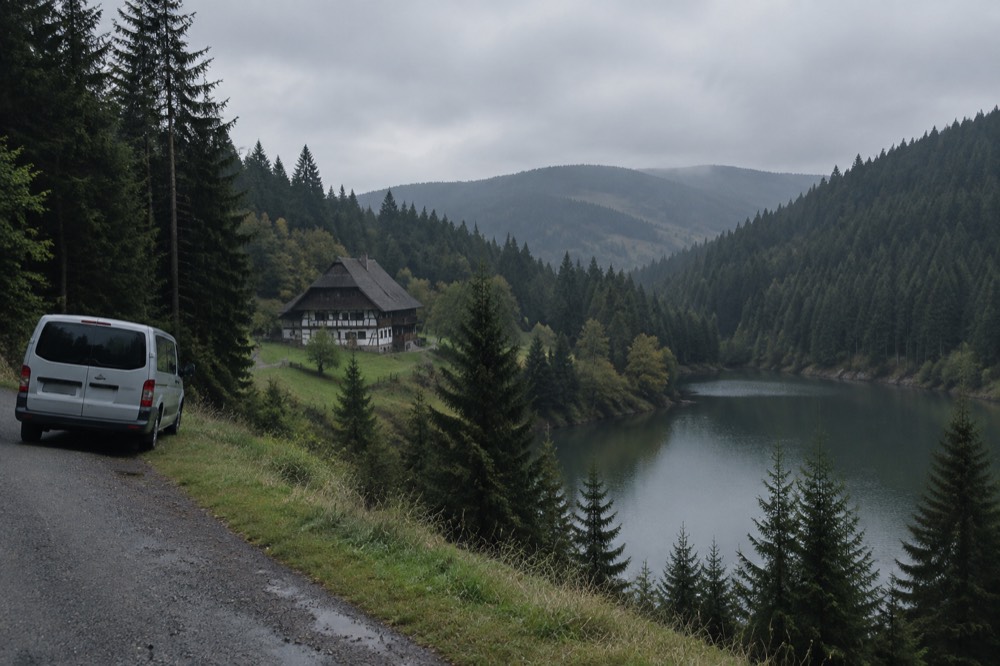 Minibus parked on a forest road above a lake surrounded by fir-covered slopes