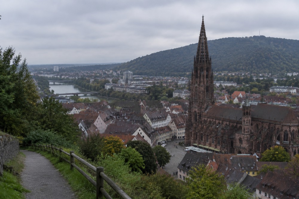 Gabled rooftops and a sandstone tower seen from a green hillside path above the city