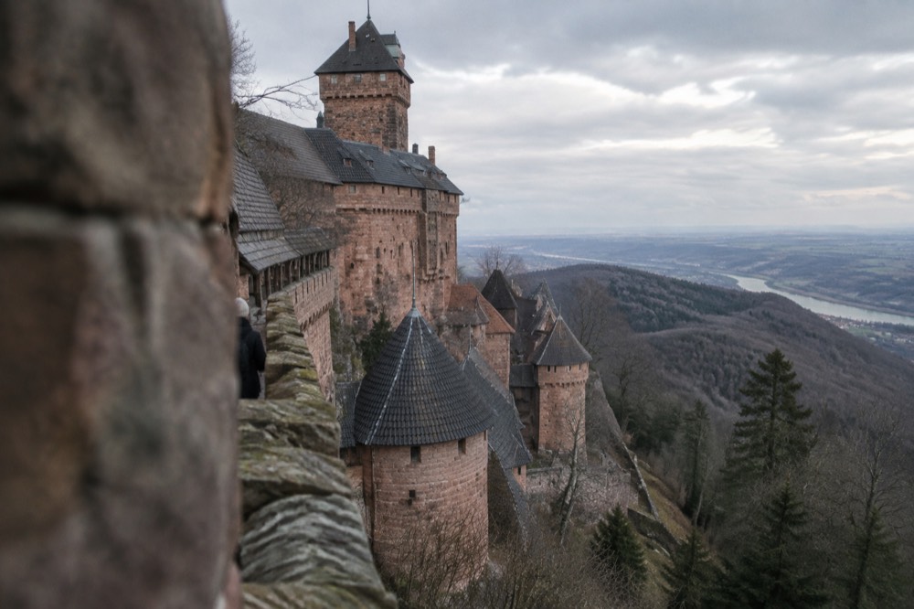 A medieval hilltop castle with round towers and pink sandstone walls above forested hills