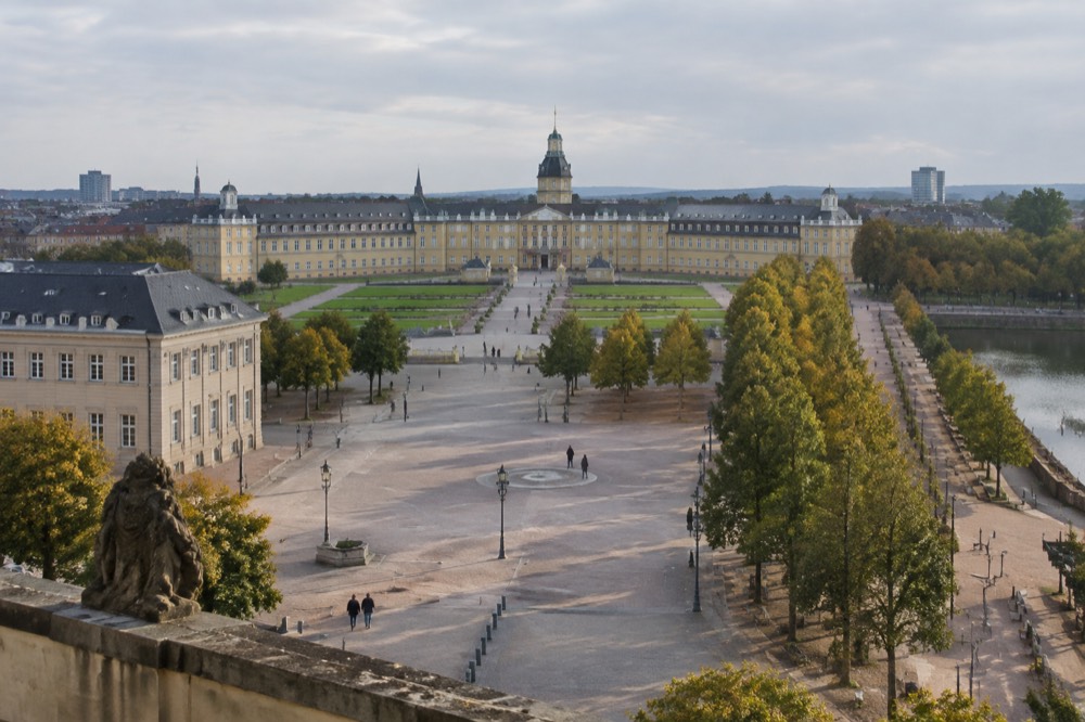 A wide German city square with a stone palace facade and tree-lined avenues under a clear sky