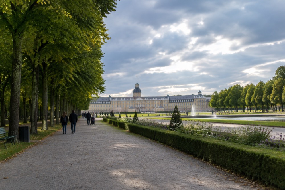 A long baroque palace garden with gravel paths, trimmed hedges and strollers under tall trees