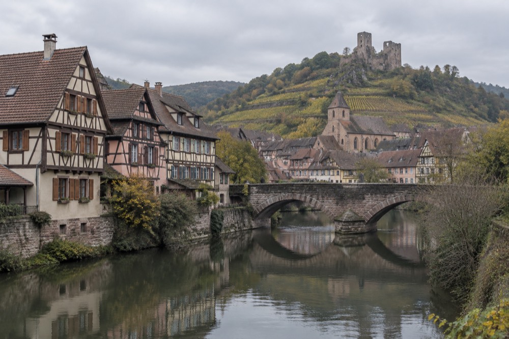 A stone bridge over a stream with half-timbered houses reflected in the water
