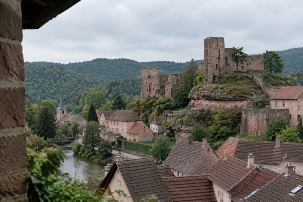 Ruined sandstone castle walls and terraces above the village roofs with forested hills beyond