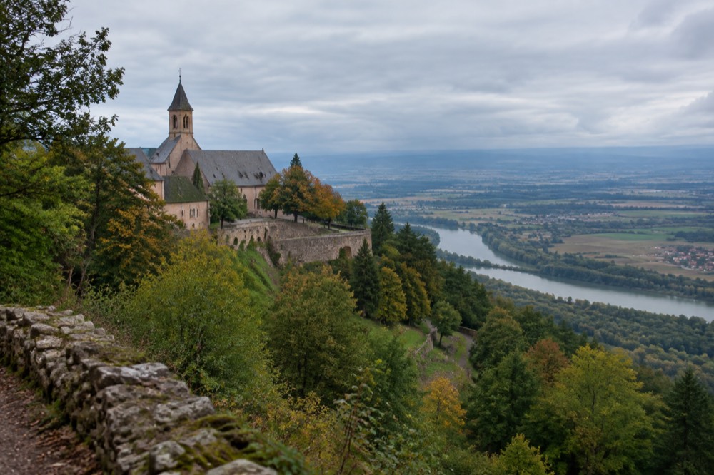 A mountaintop abbey with pale walls and a bell tower seen from a forest road on a ridge in the Vosges foothills