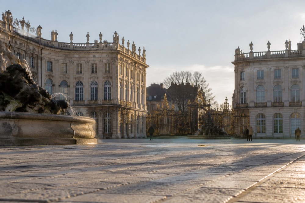A grand classical square with gilded wrought iron gates and fountains under a clear sky