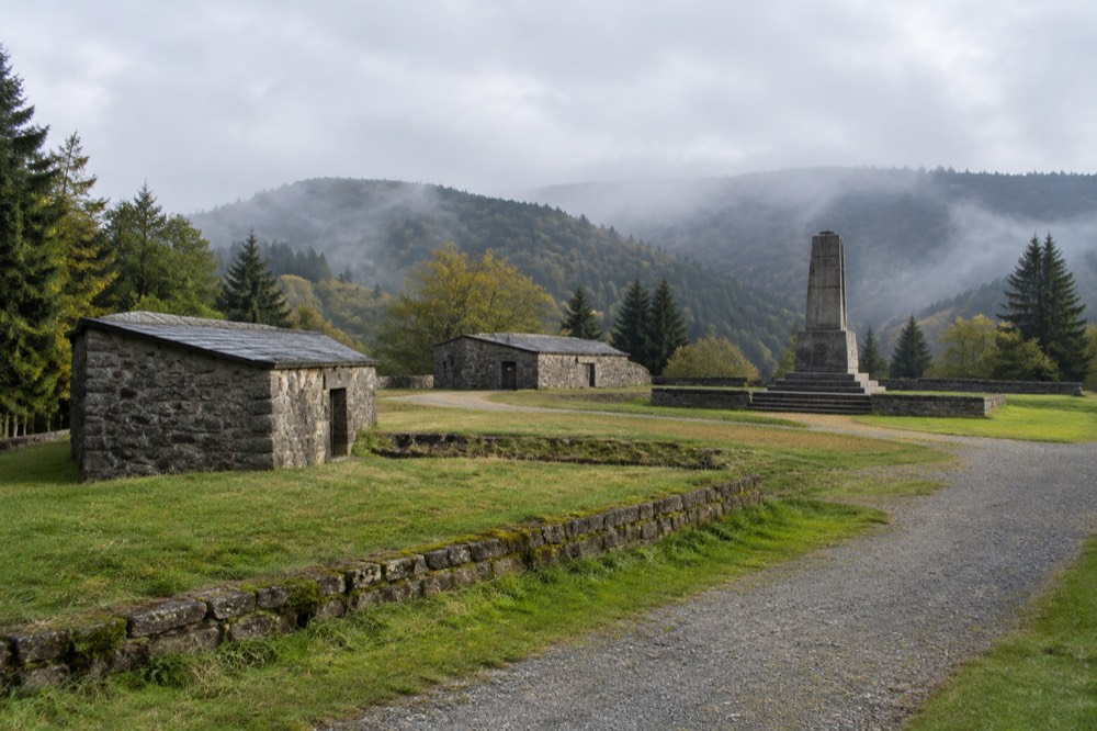 A wide view of a hilltop clearing with low stone buildings and a monument in the Vosges mountains