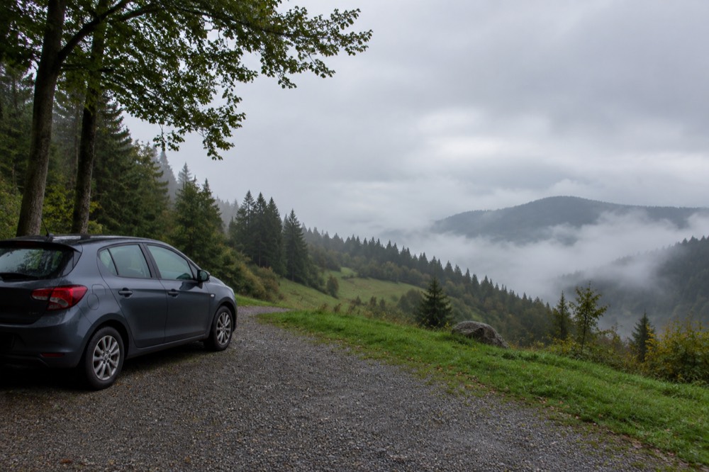 A hire car parked at a forested roadside viewpoint with mist over a valley