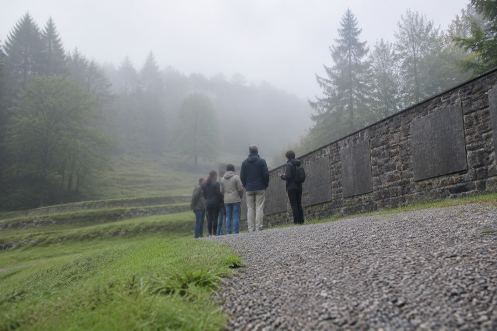 A small group of visitors standing quietly beside a guide at an outdoor memorial wall