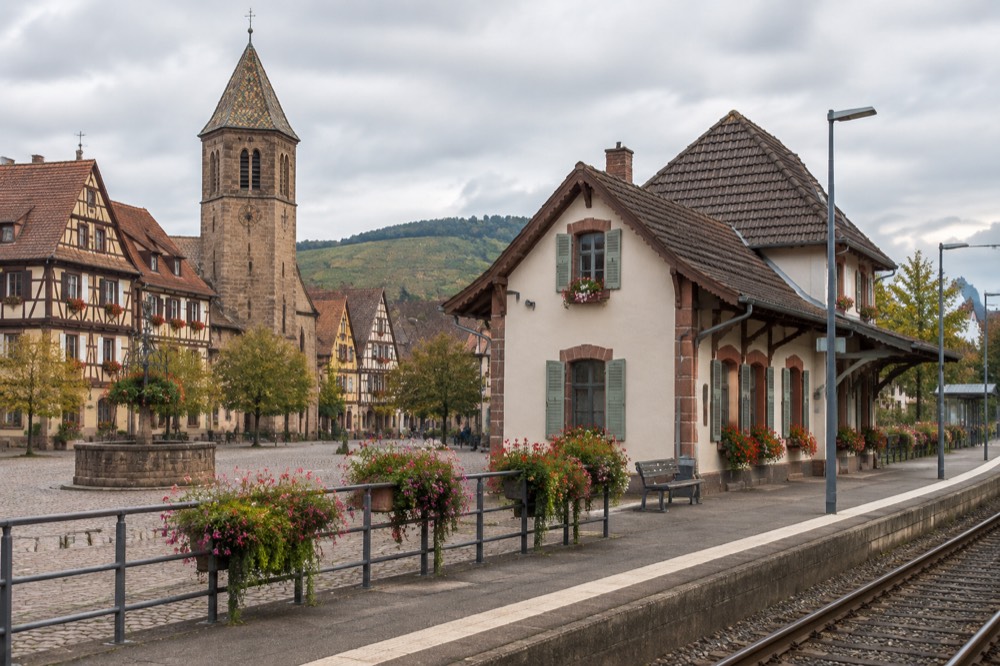 A regional train platform beside a small Alsatian station building with flower displays