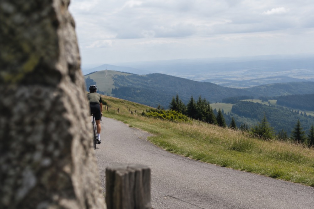 A narrow ridge road curving between grassy Vosges meadows with views over a wide plain