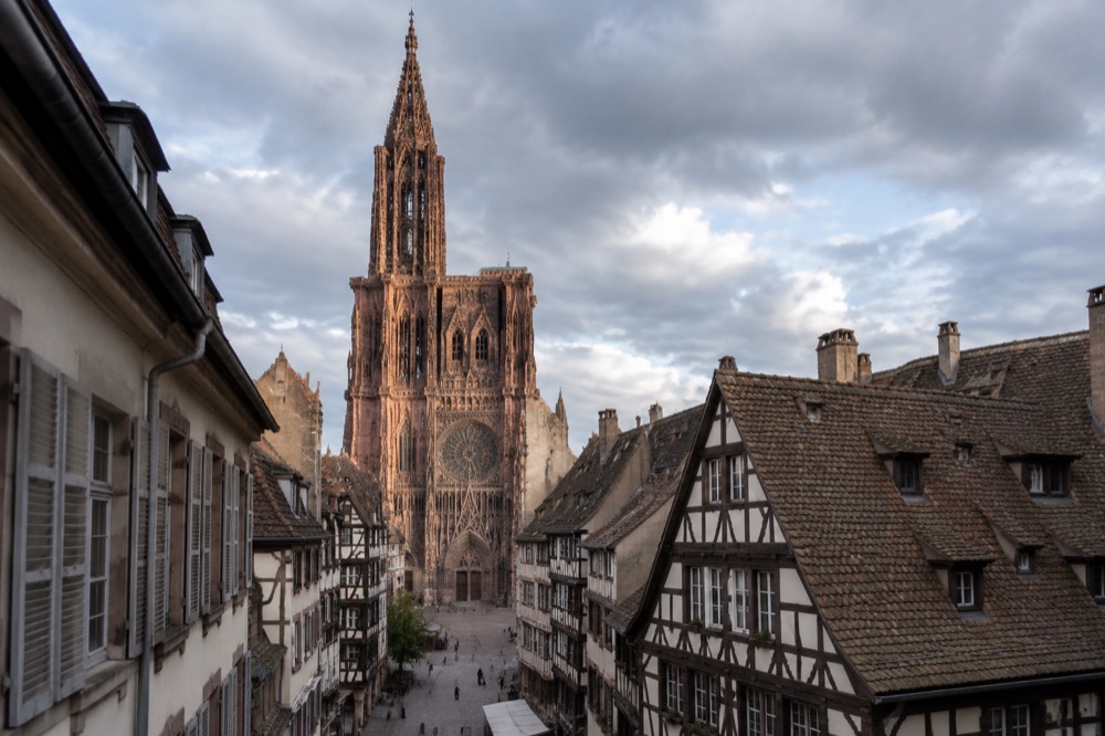 A pink sandstone Gothic cathedral rising above rooftops seen from a narrow old street