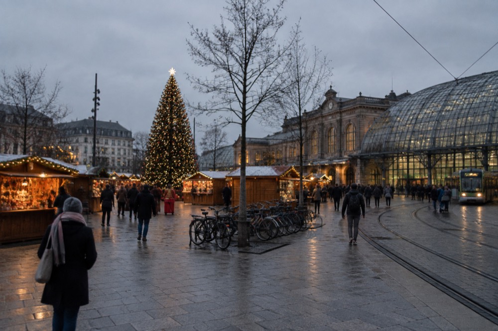 A Christmas market square at dusk with illuminated wooden chalets and a large decorated tree
