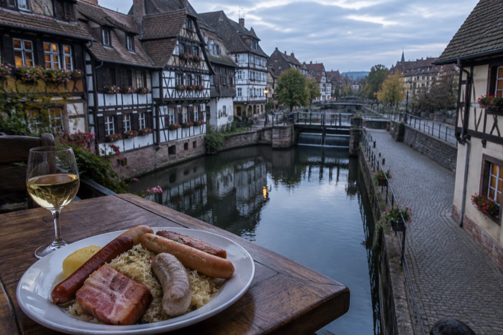 A plate of choucroute garnie with sauerkraut, sausages and pork beside a glass of Alsatian white wine on a wooden winstub table