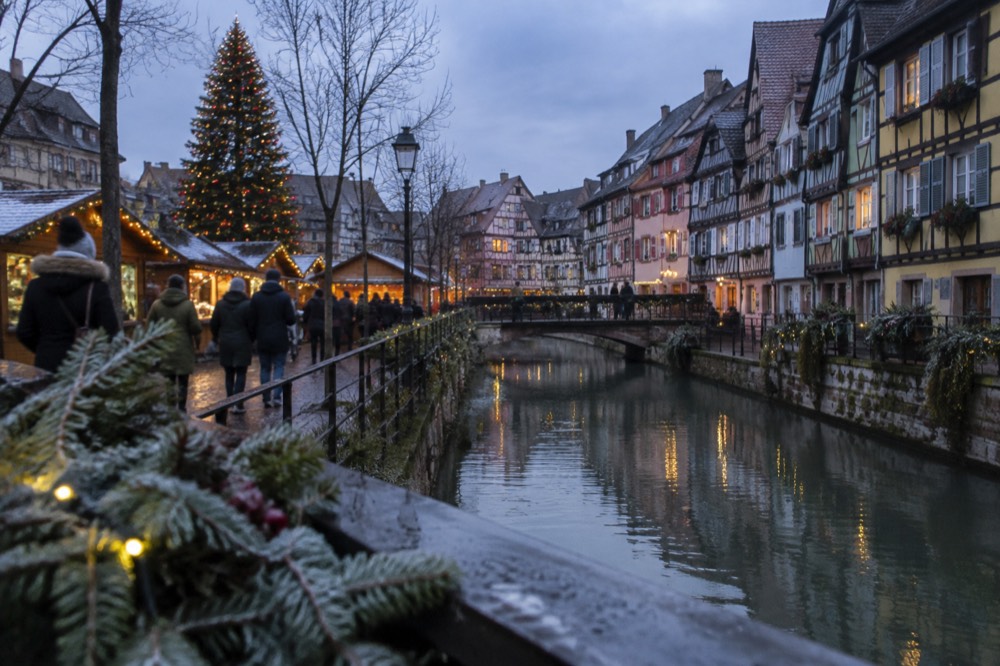 A Christmas market square with wooden chalets and a large decorated tree at dusk
